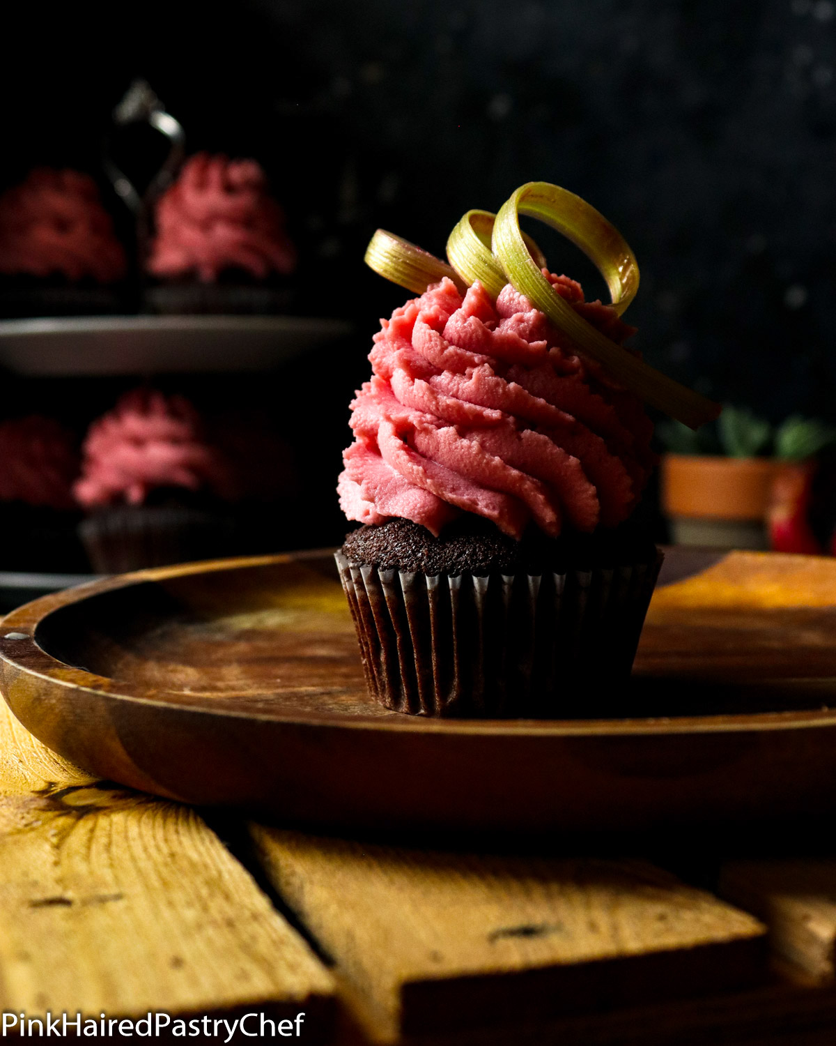 Chocolate Cupcake on a wooden plate on top of a wooden board. Hot Pink Rhubarb Whipped Ganache piped high on top with star nozzle. Rhubarb curl garnish on top.