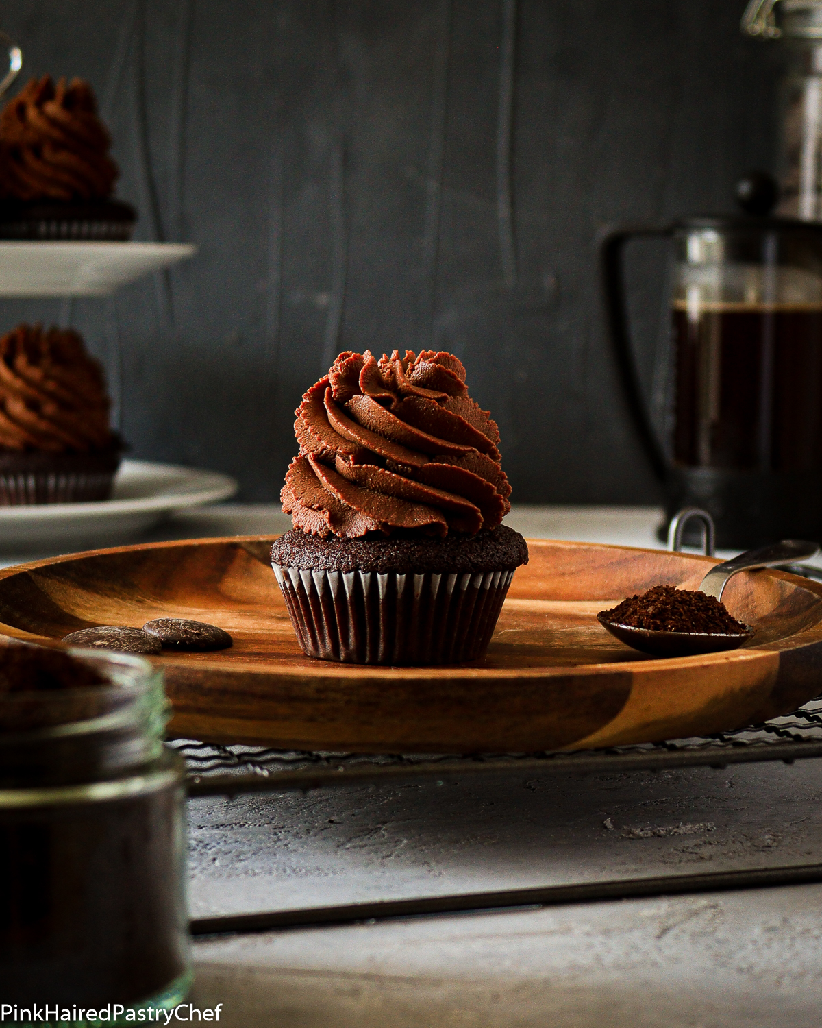 Chocolate Cupcake with Coffee Dark Chocolate Whipped Ganache piped high on top with star nozzle, Cupcake is on a wooden plate on top of a black wire rack, More cupcakes on cupcake stand in background along with french press full of coffee. Small jar of ground coffee to the side for decoration
