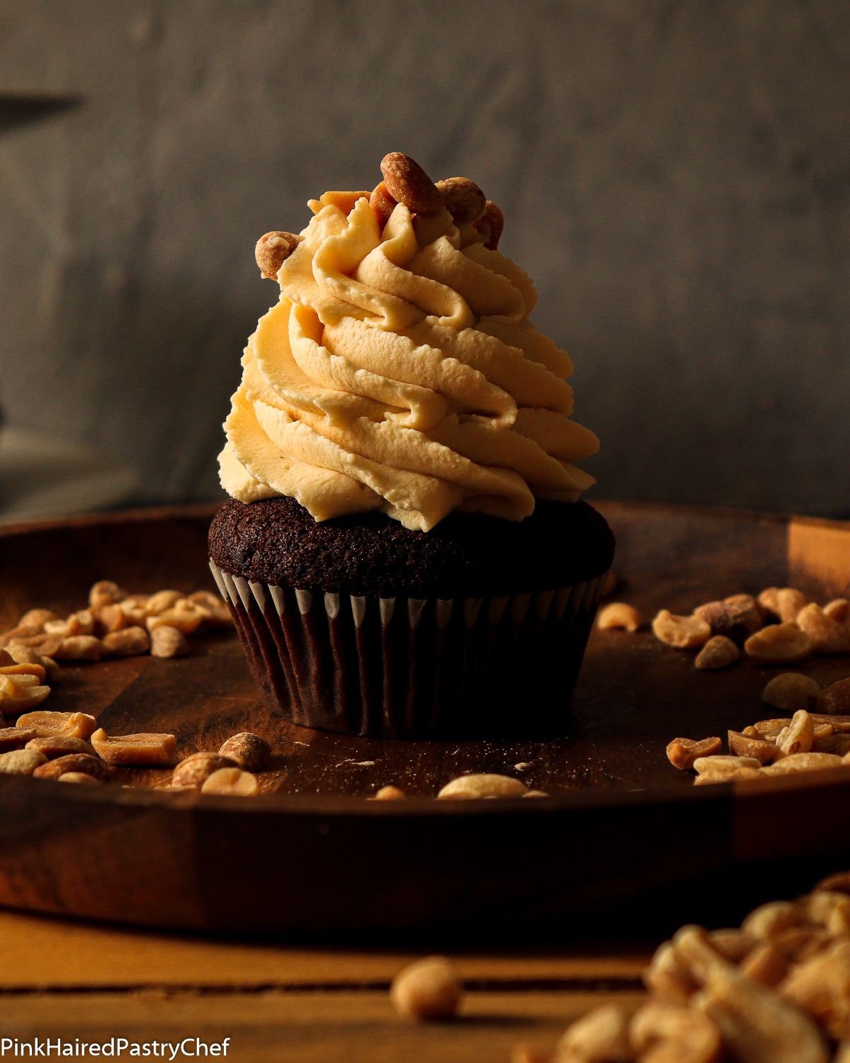 Chocolate Cupcake on top of a wooden plate, topped with Peanut Butter White Chocolate Whipped Ganache piped high on top with star nozzle. Topped with roasted peanut.