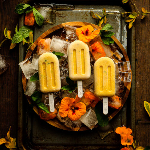 3 Malibu Mango Yellow coloured Frozen Pops laying flat on a wooden tray covered with ice cubes and fresh flowers